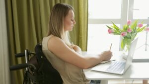 Woman in wheelchair writing on paper and looking at computer.